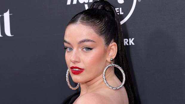 Sixtine smiles over her shoulder, wearing a black one-shoulder dress, oversized silver hoops and a red lip.